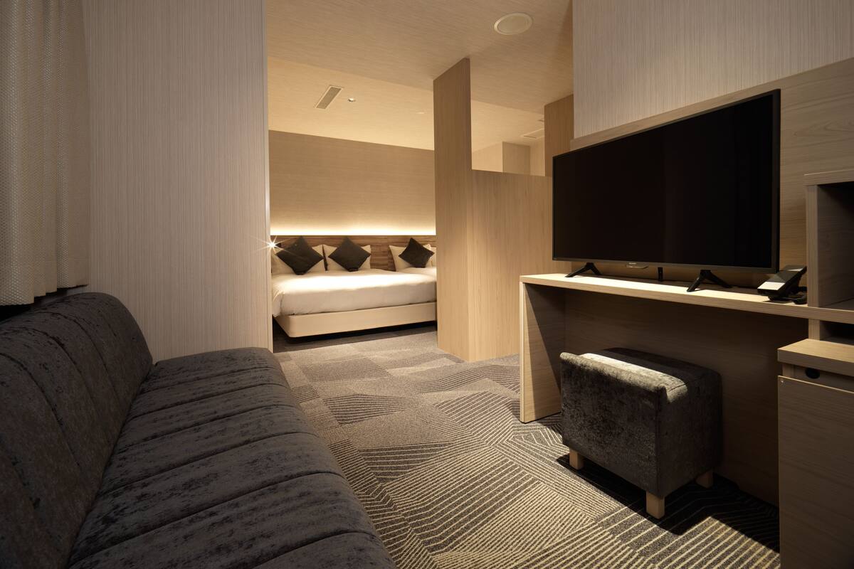 A modern hotel room with a sofa, a desk with a flat-screen TV, a bed with a headboard, and a carpeted floor.