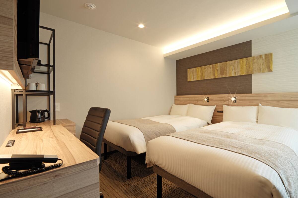 A hotel room with two beds, a desk with a telephone, a chair, a coffee maker, and a shelf with cups.