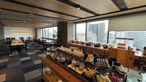Uma área de refeições moderna com piso xadrez, mesas e cadeiras de madeira. Há um balcão de buffet com diversos alimentos e uma placa com a inscrição "Hareus".