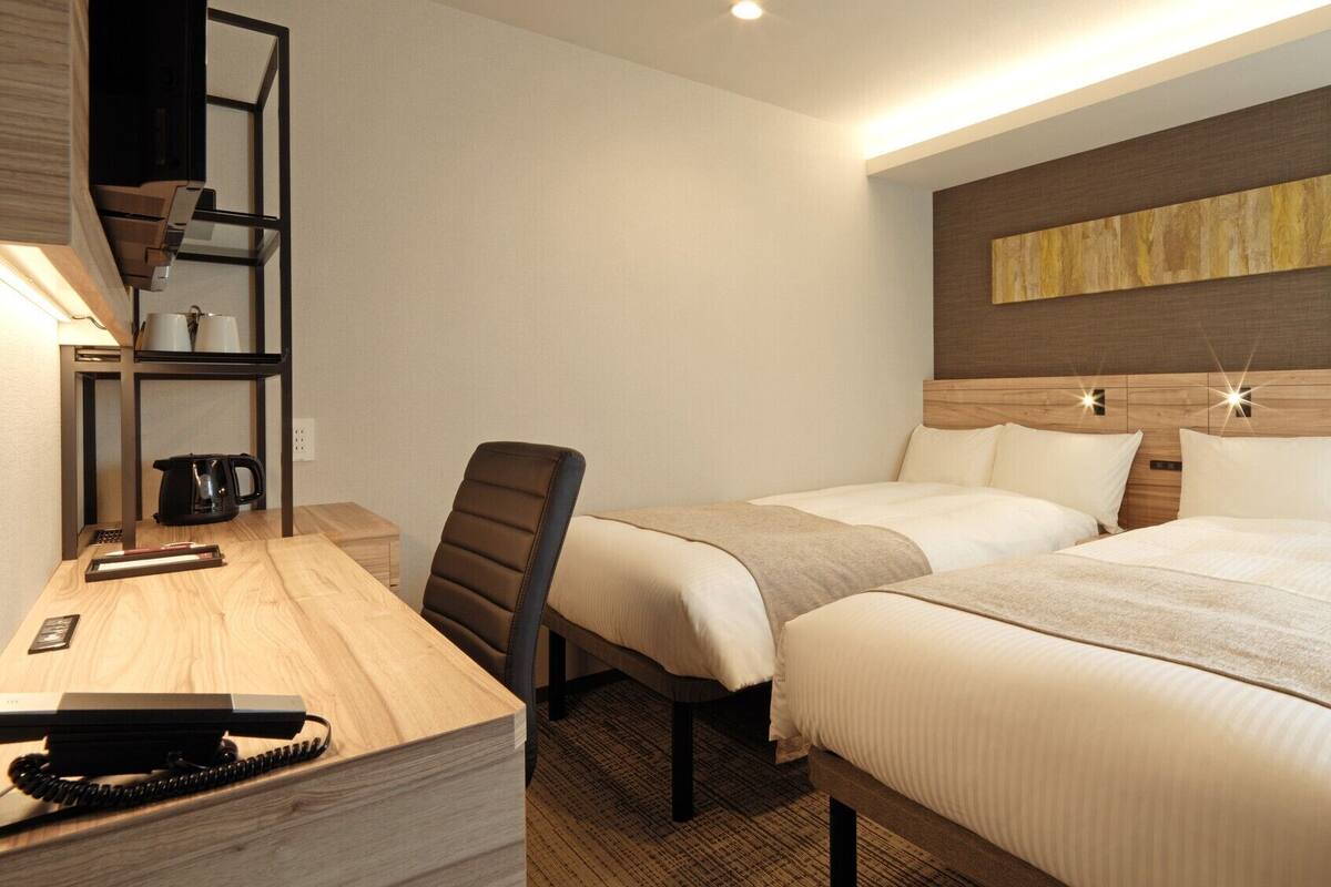 A hotel room with a desk, chair, two beds, a coffee maker, and a wall art piece.