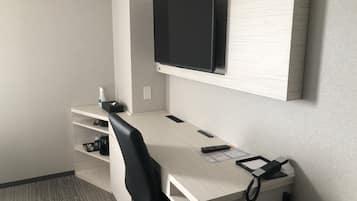 A hotel room with a wooden desk, a black office chair, a mounted flat-screen TV, a telephone, and a shelf with a bottle and tissues.
