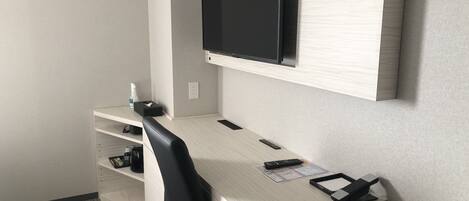 A hotel room with a wooden desk, a black office chair, a mounted flat-screen TV, a telephone, and a shelf with a bottle and tissues.