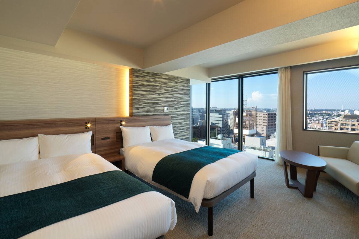A hotel room with two beds, a sofa, a small table, and a large window offering a city view.