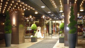 Entrada de hotel com porta de vidro, um manequim vestido de noiva e luzes decorativas.