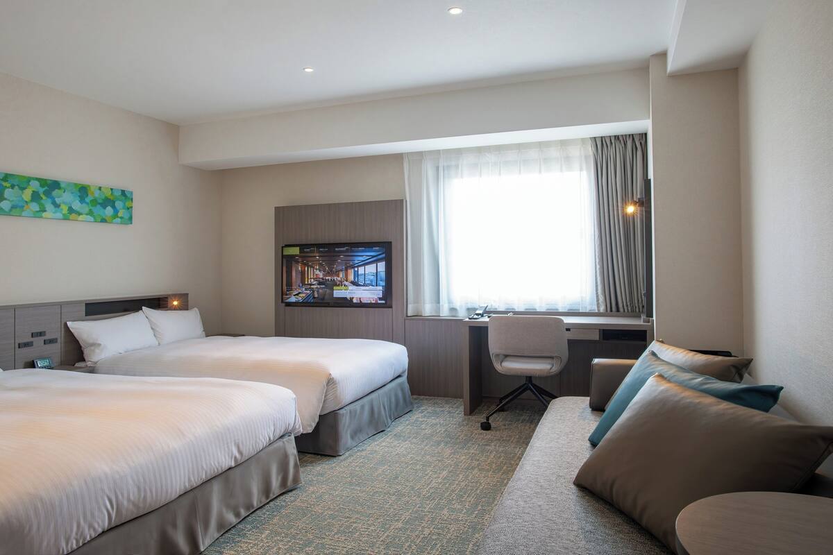 A hotel room with two beds, a desk, a chair, and a large window with curtains.