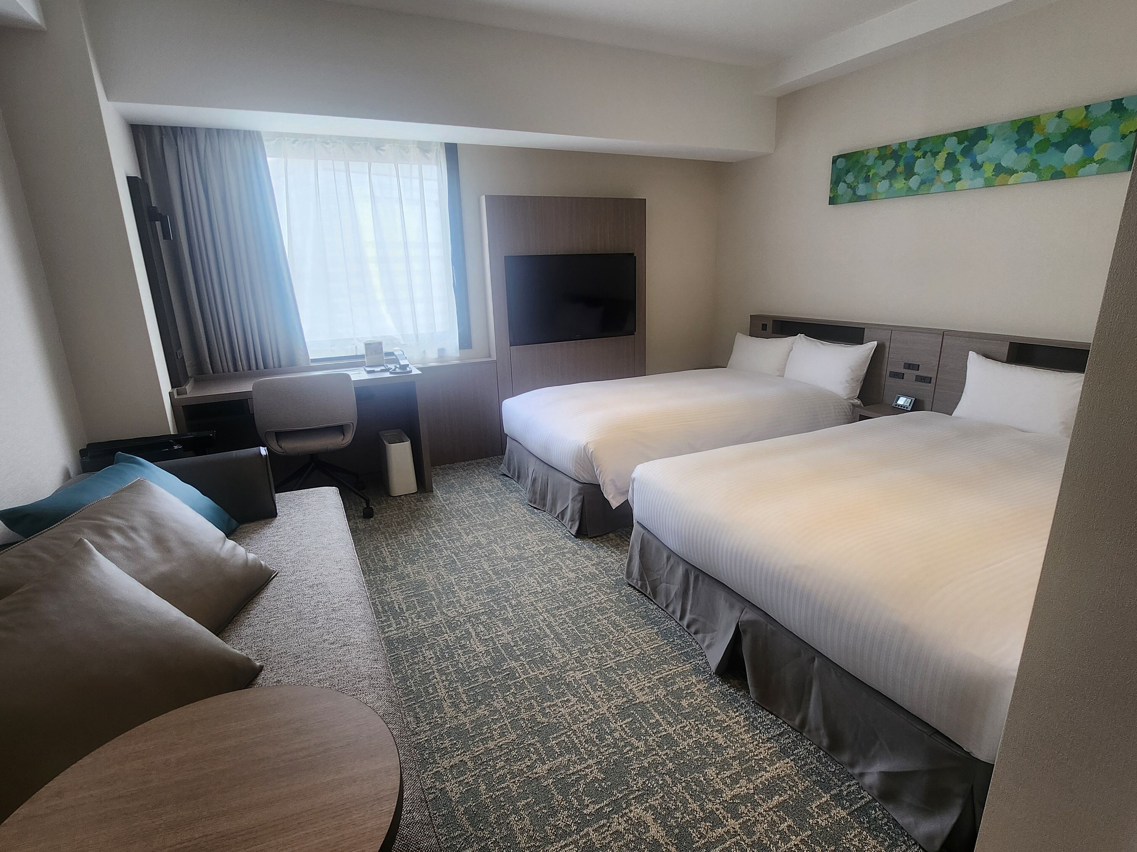 Ein Hotelzimmer mit zwei Betten, einem Schreibtisch, einem Stuhl, einem Fernseher und einem Fenster mit Vorhängen.