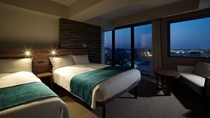 Quarto de hotel com duas camas, vista da cidade à noite e varanda com mesa e cadeira.