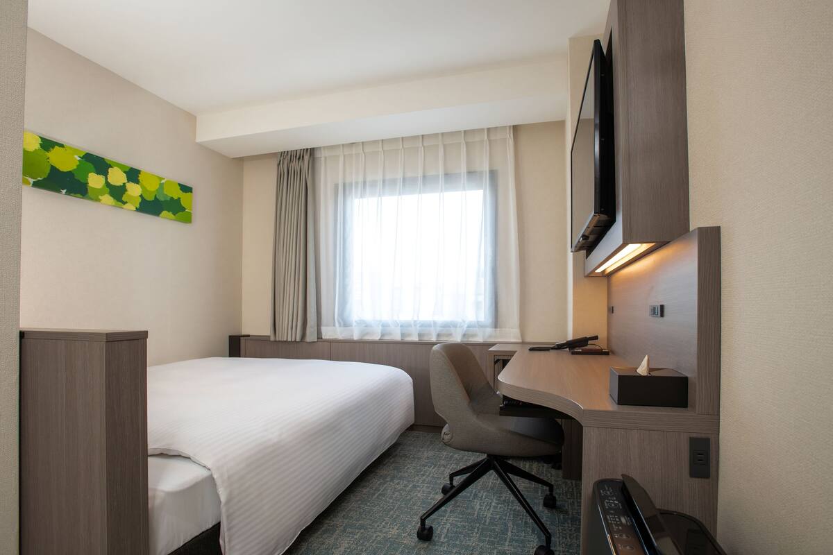A hotel room with a bed, a desk with a chair, a television, and a window with curtains.
