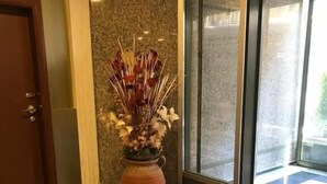 Eine Hotelhalle mit Schachbrettmuster-Teppich, einem roten Teppich mit dem Namen des Hotels, einer großen Vase mit Trockenblumen und einer Tür links.