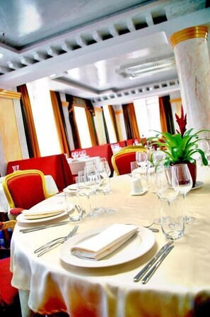 A dining table set with plates, cutlery, and wine glasses, surrounded by red chairs in a well-lit room with a ceiling design. - Actor Hotel Budapest  (Budapest)