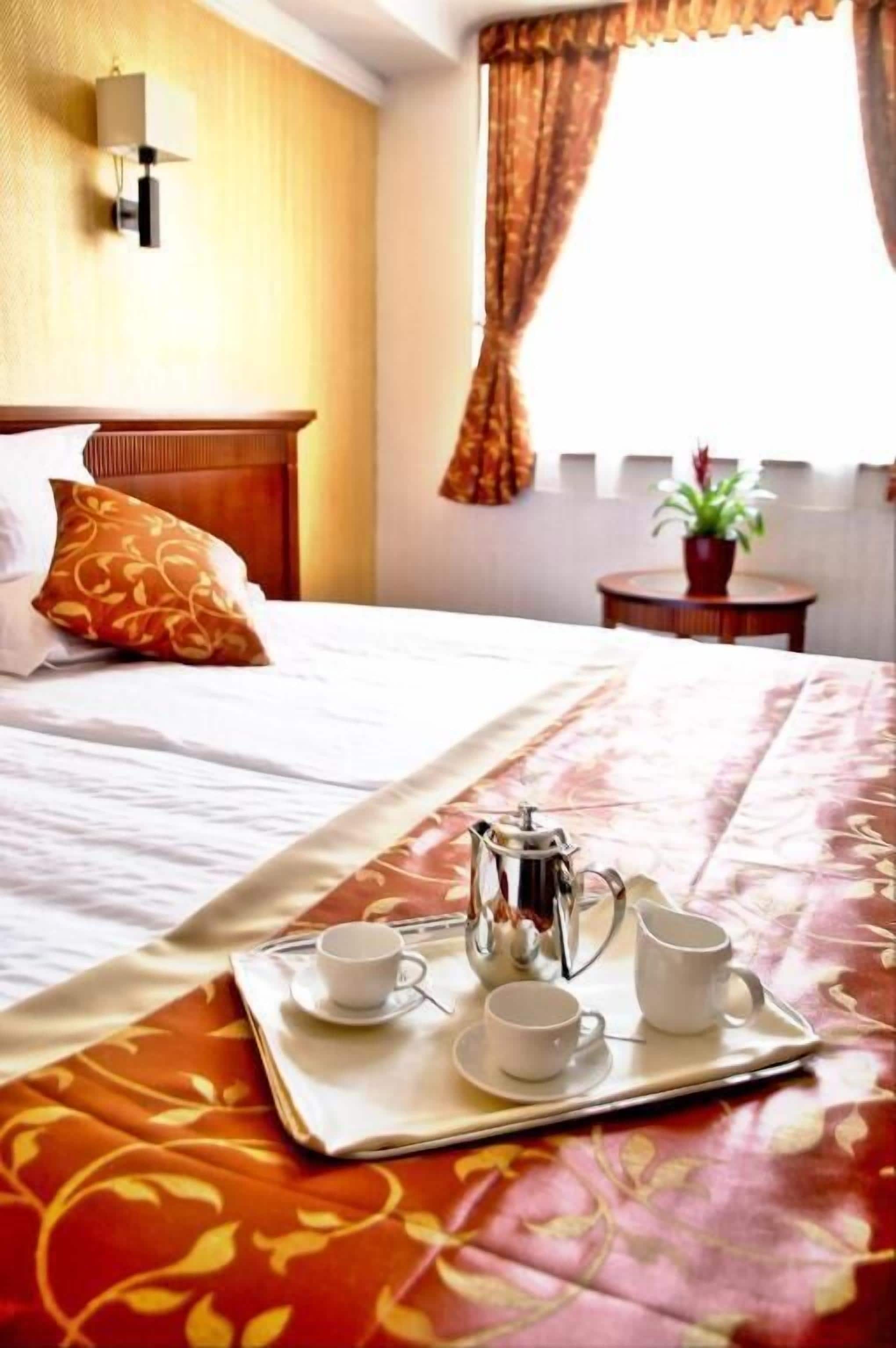 Habitación de hotel con una cama bien hecha, una bandeja con una cafetera y tazas, y una planta en maceta sobre una mesita.