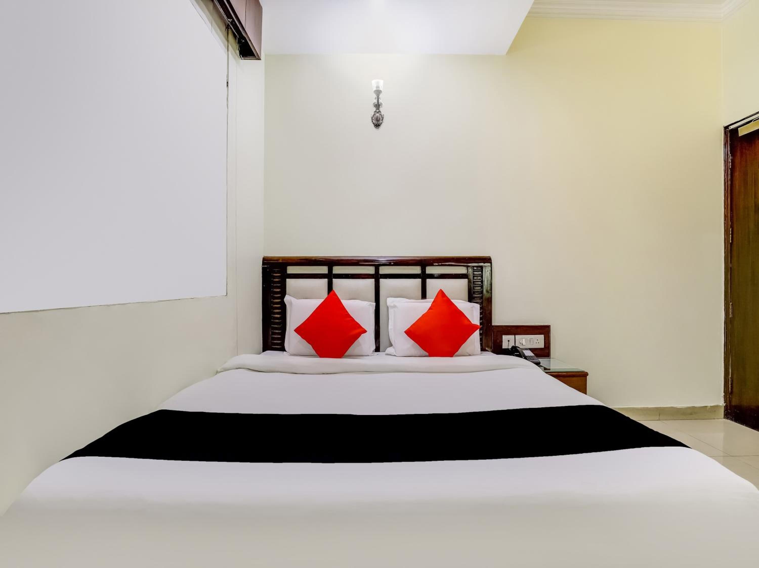 A neatly made bed with white bedding and two red pillows, a bedside table with a phone, and a wall-mounted light fixture.
