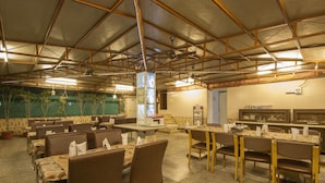A spacious restaurant with multiple tables and chairs, a high ceiling with exposed beams, and a kitchen area visible in the background. - Hotel Grand Park Inn (New Delhi)