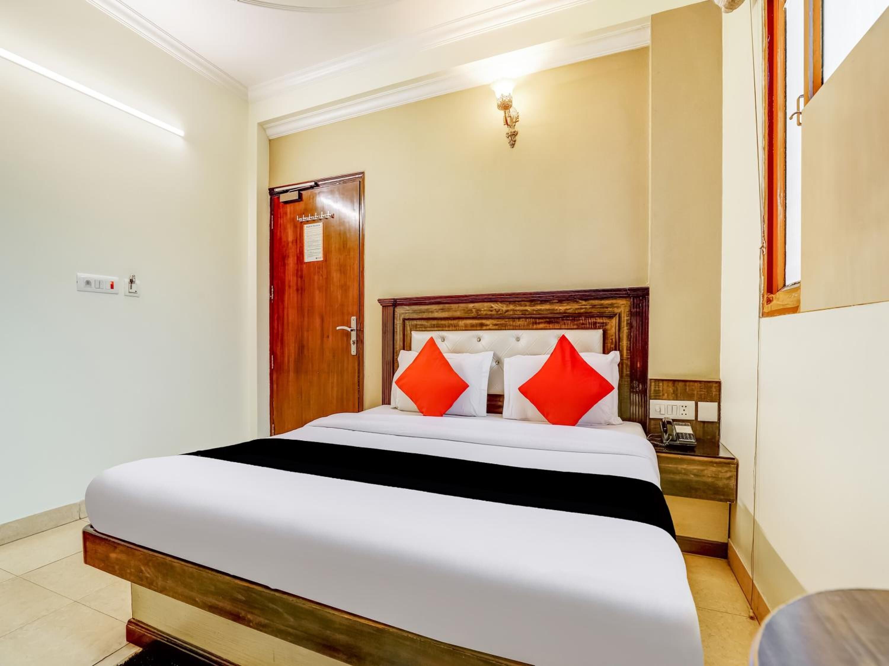 A hotel room with a bed, a wooden door, a nightstand, and a wall-mounted light fixture.