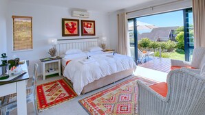 Un dormitorio con una cama grande, un escritorio con silla, un balcón con mobiliario de mimbre y obras de arte en las paredes.