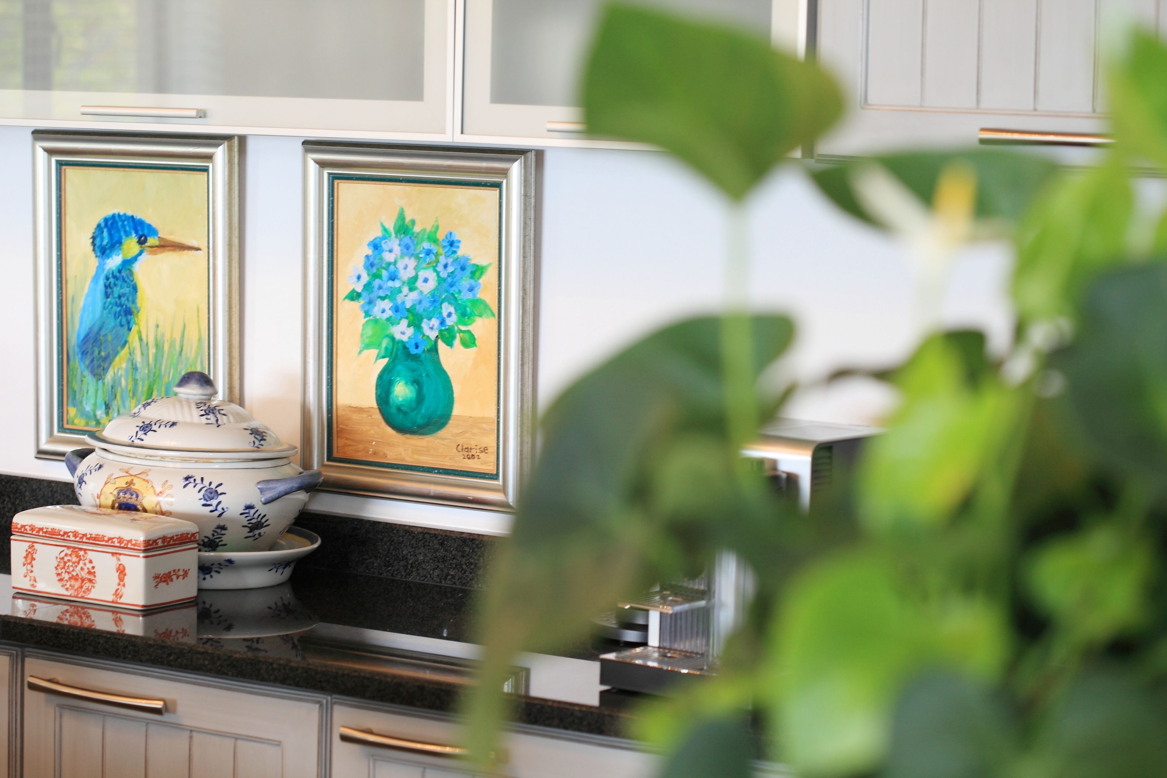 A kitchen with framed artwork, a white cabinet, and a plant in the foreground.
