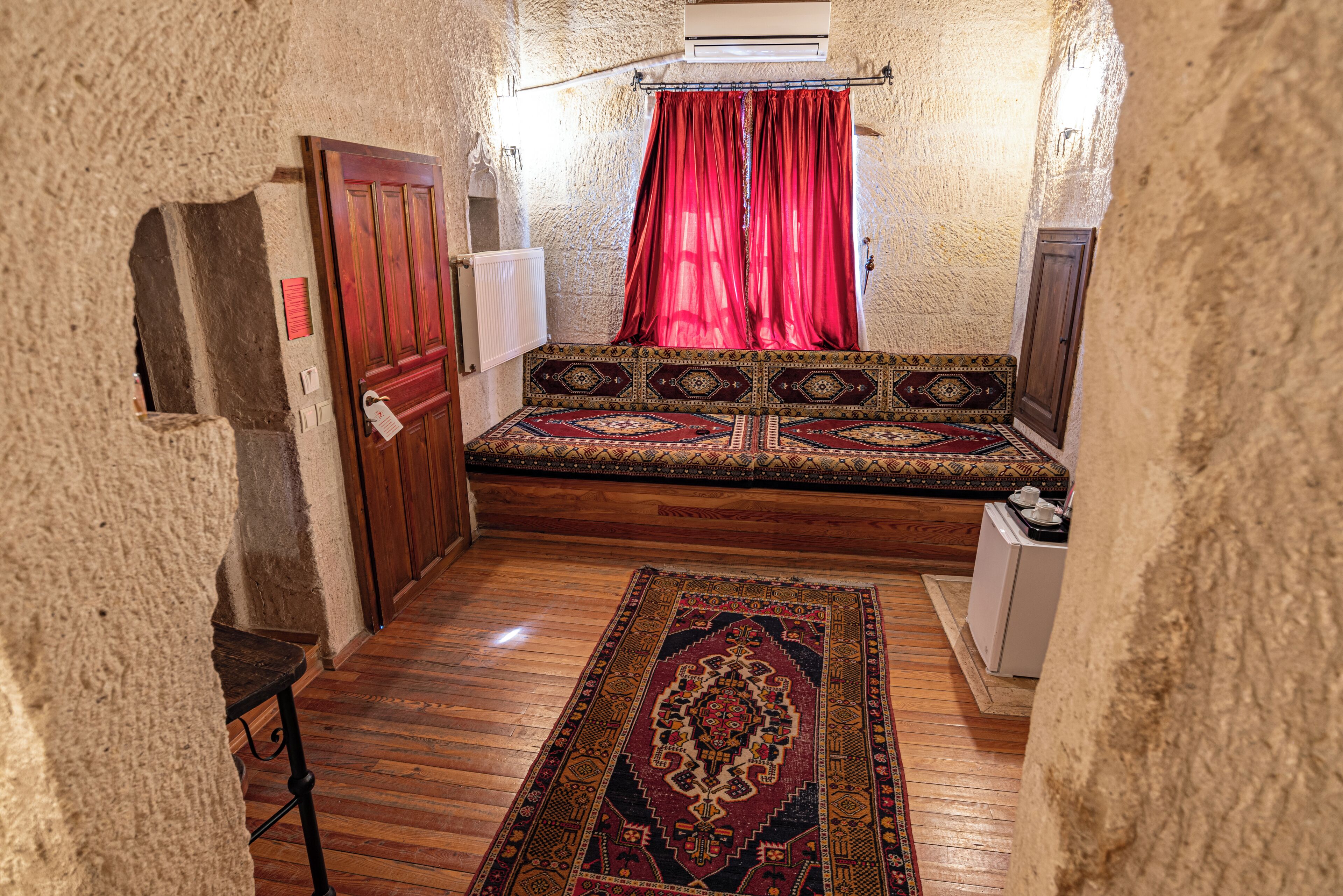 A narrow room with a wooden floor, a patterned rug, a bench with a cushion, a red curtain, and a small cabinet.