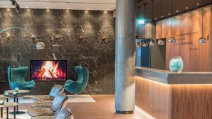 Een moderne lobby met een grote televisie waarop een haard te zien is, lederen zitplaatsen, een houten receptiebalie en een marmeren wand met klokken.