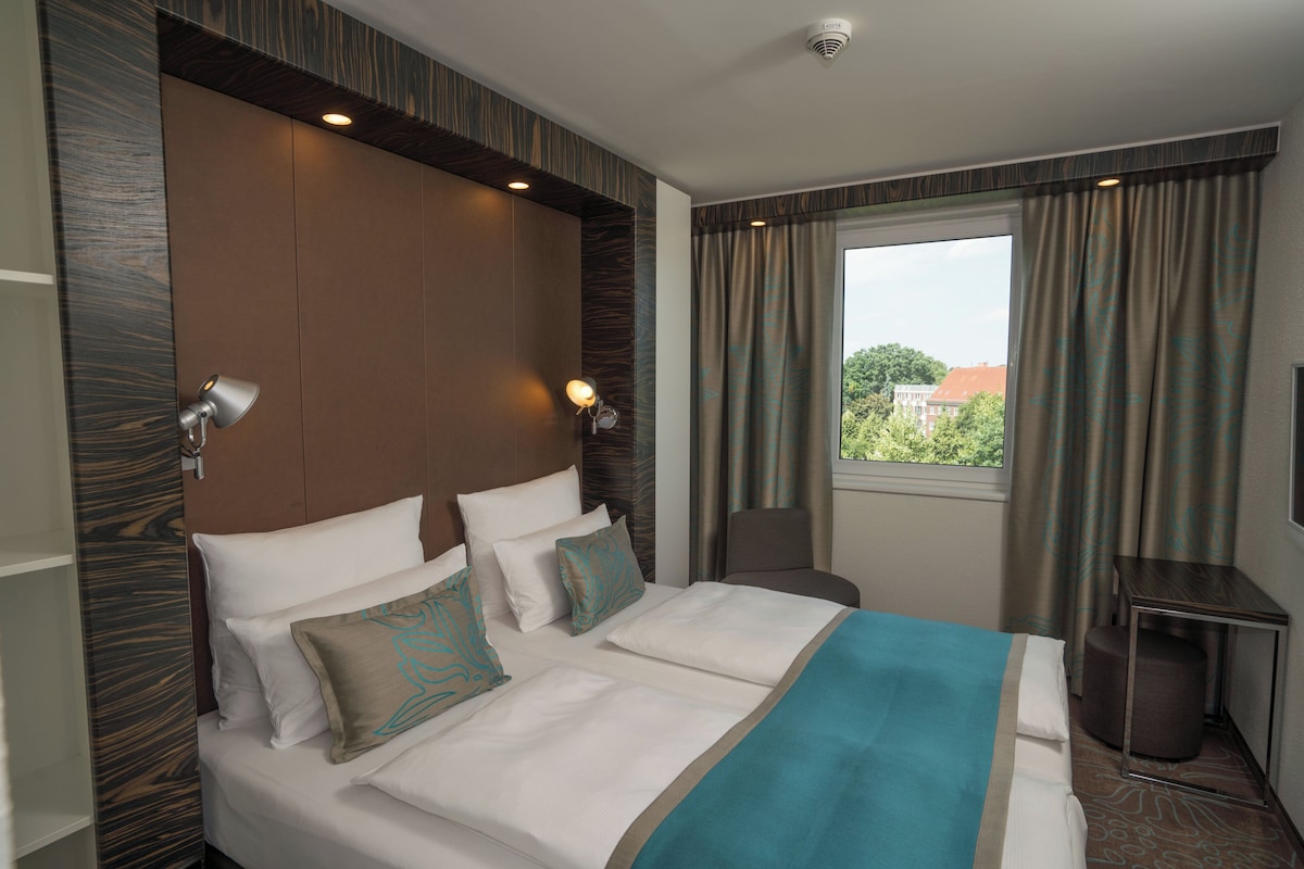 A hotel room with a large bed, two bedside lamps, a window with curtains, and a view of greenery outside.
