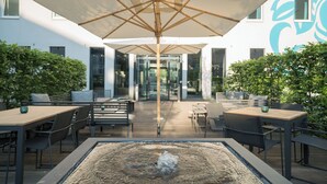 Een buitenpatio met tafels en stoelen, een centraal vuurkorf en een grote parasol voor schaduw.