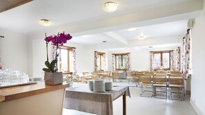 Un espace repas comprenant plusieurs tables et chaises, une table centrale sur laquelle sont empilées des assiettes, et un pot de fleurs orné de fleurs violettes.