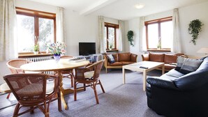Un salon comprenant un coin repas, un canapé, un téléviseur et de grandes fenêtres avec des rideaux.