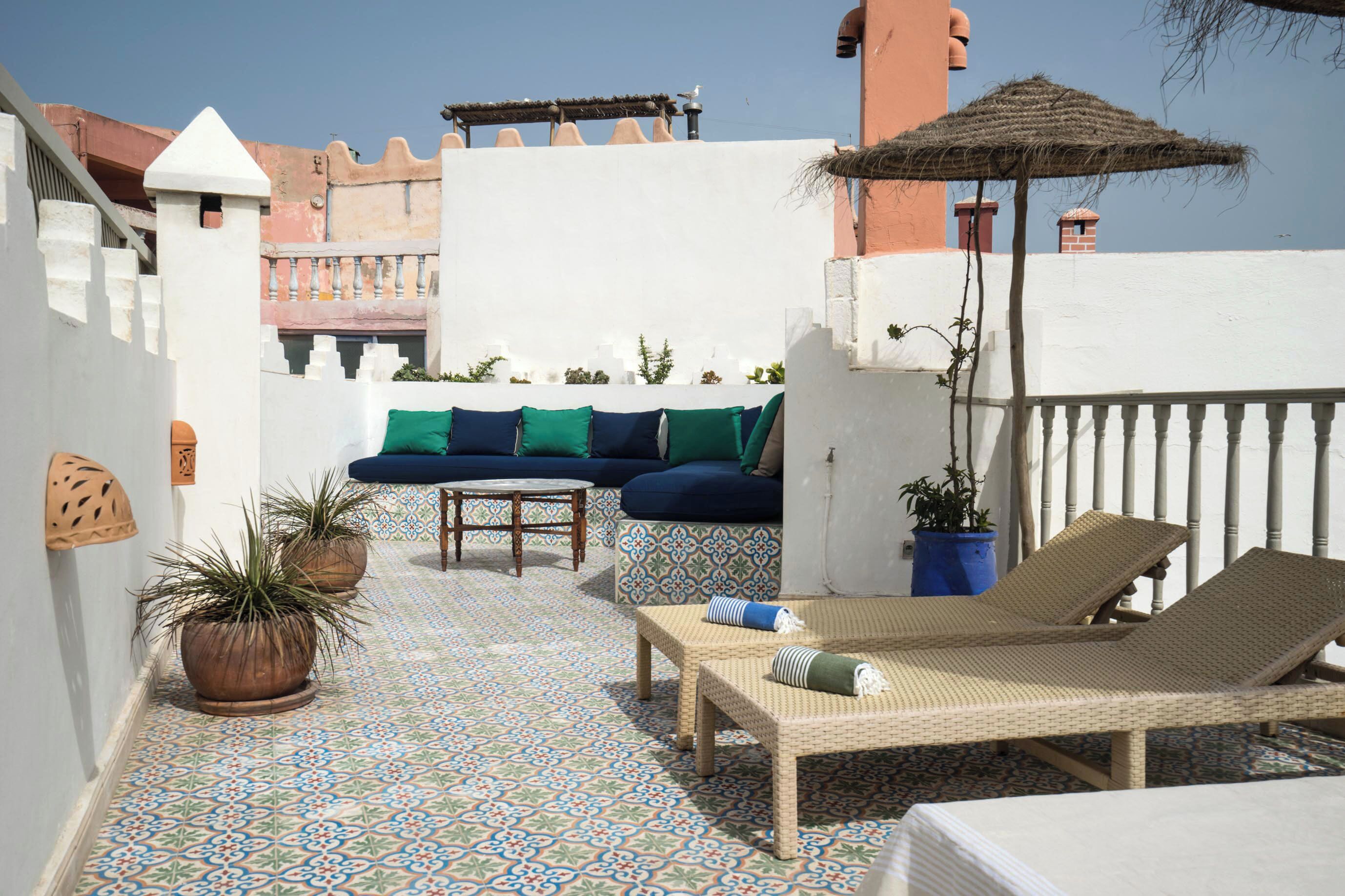 A rooftop terrace with a patterned floor, a sofa with cushions, a lounge chair, and potted plants.