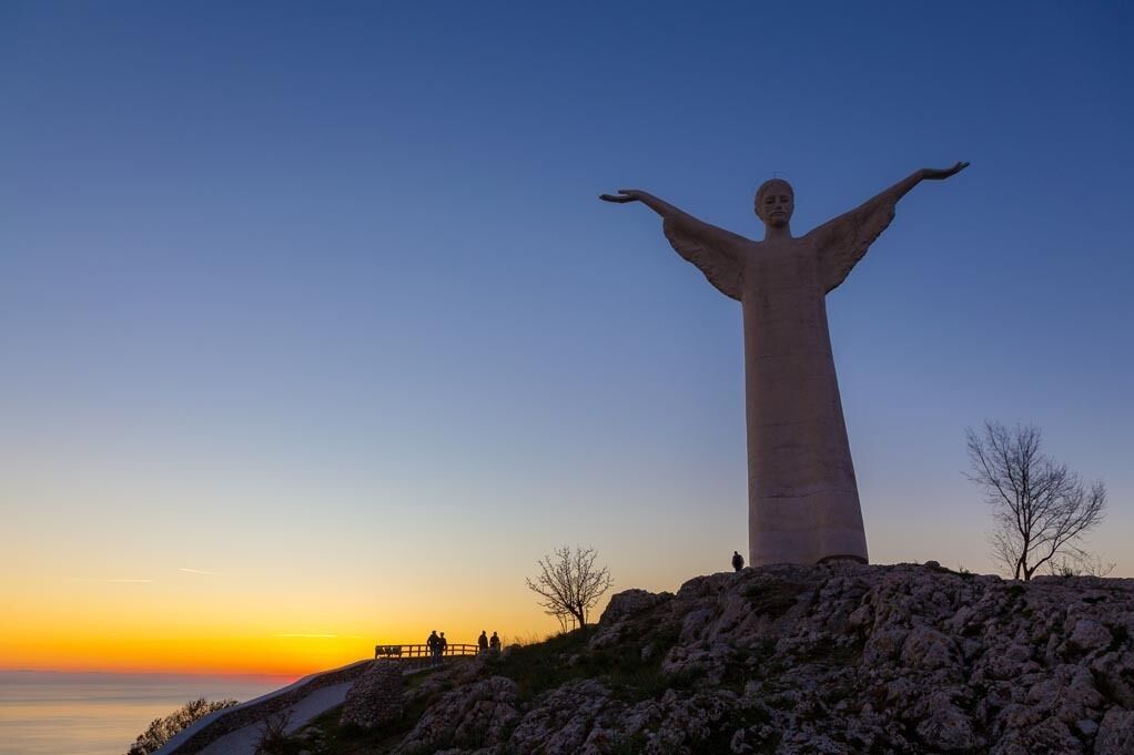 A large statue with outstretched arms on a rocky hill overlooking a body of water during sunset.