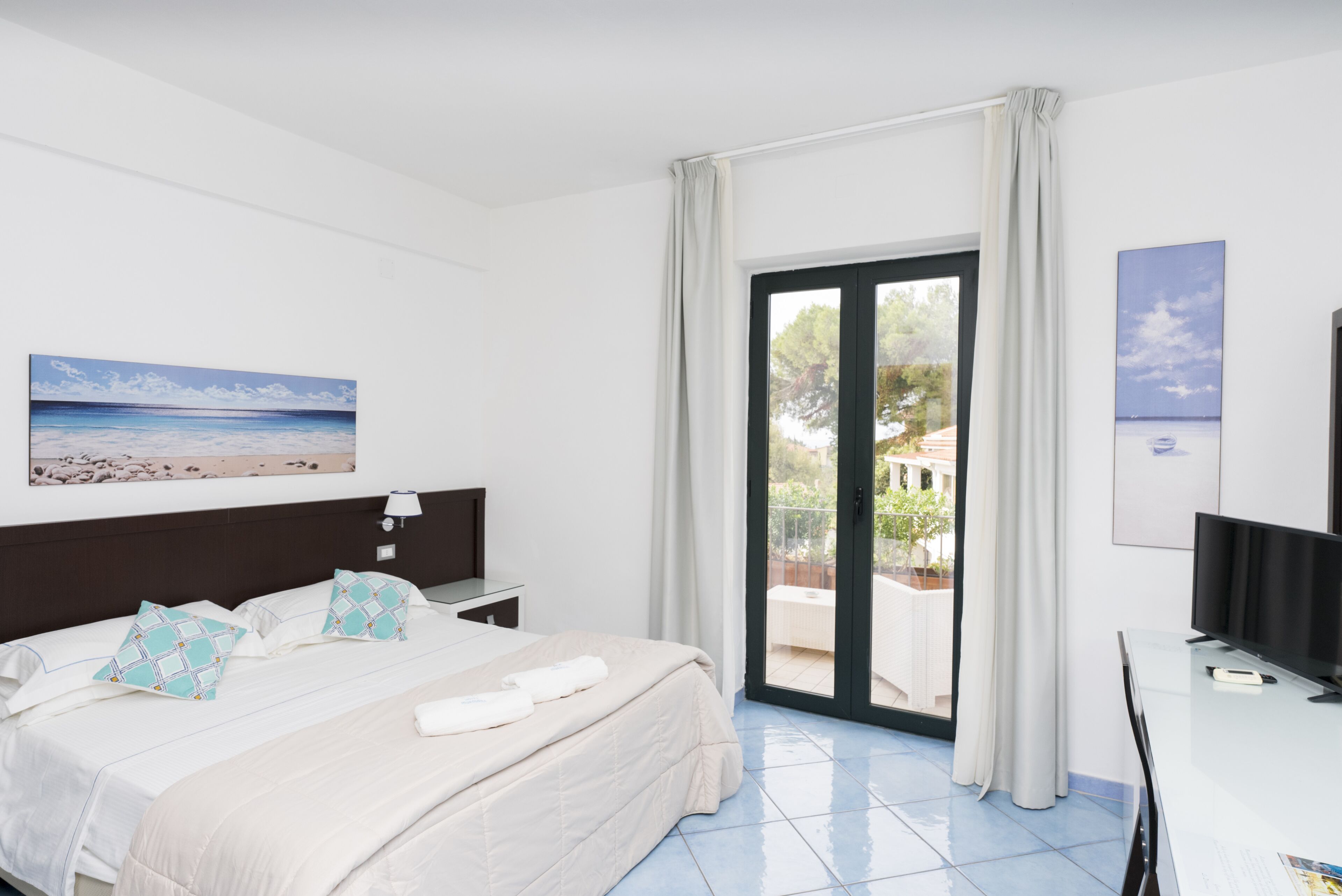 A hotel room with a bed, a desk, a TV, and a balcony with a view of the sea.
