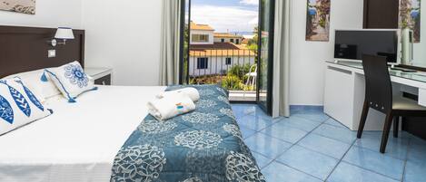 Quarto de hotel com cama, escrivaninha, cadeira e varanda com vista para edifícios e céu limpo.