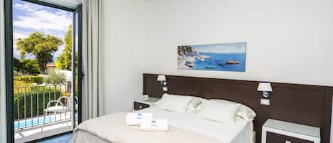 Una habitación de hotel con una cama, una mesita de noche, un escritorio, un cuadro con una playa y vistas a una piscina y zonas verdes.