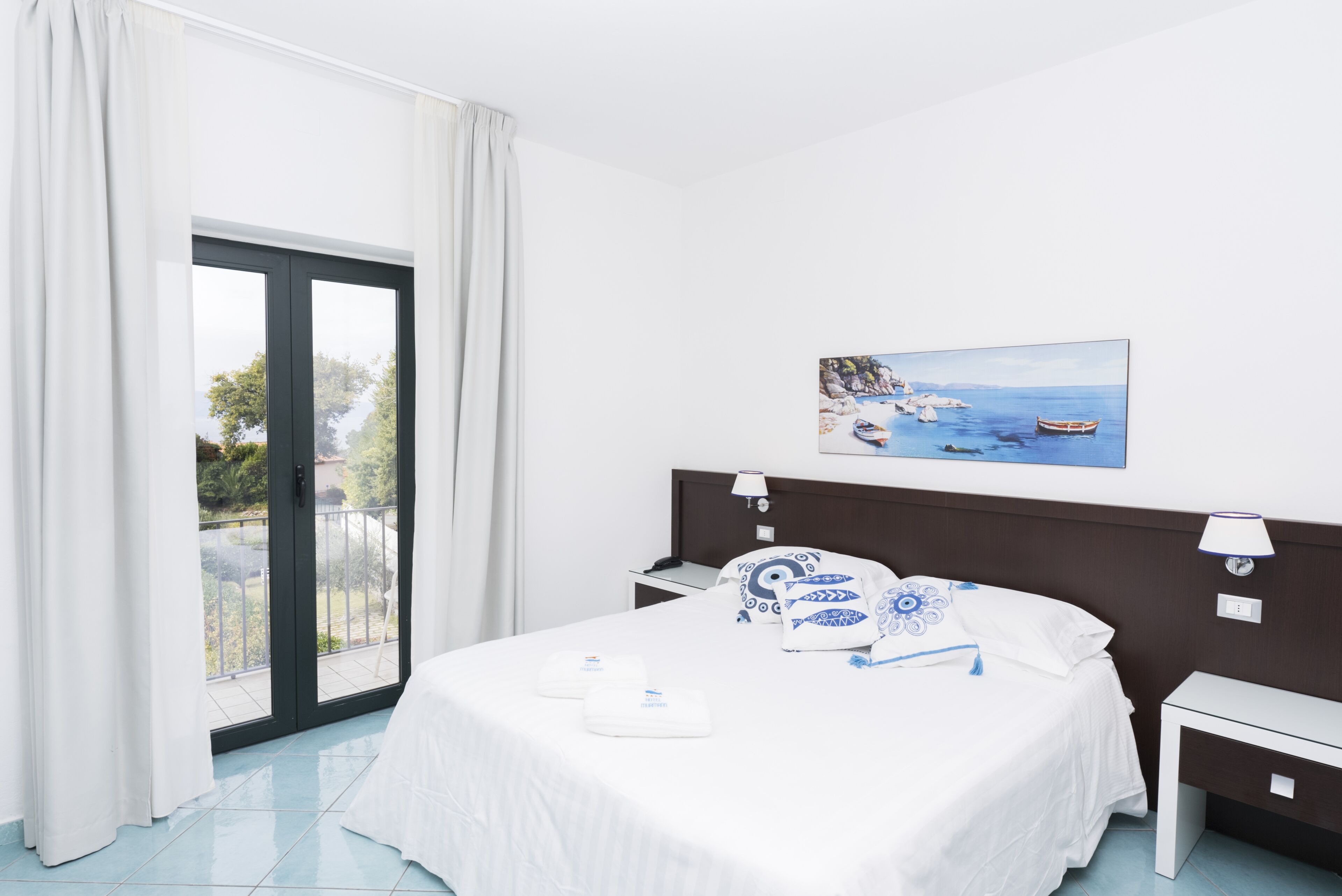 Una camera d'albergo con un letto, un comodino, un tavolino di fianco al letto, un quadro raffigurante una costa e una porta finestra scorrevole verso l'esterno.