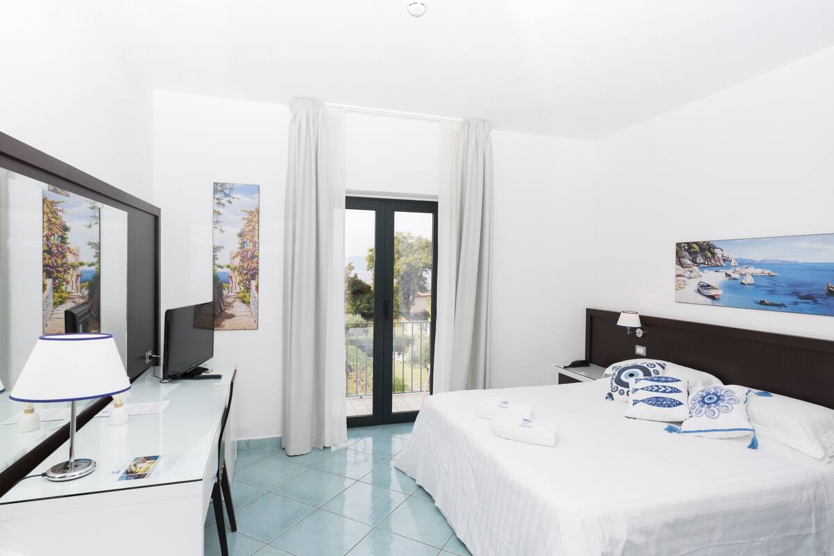 A hotel room with a bed, a desk with a lamp, a television, and a painting on the wall.