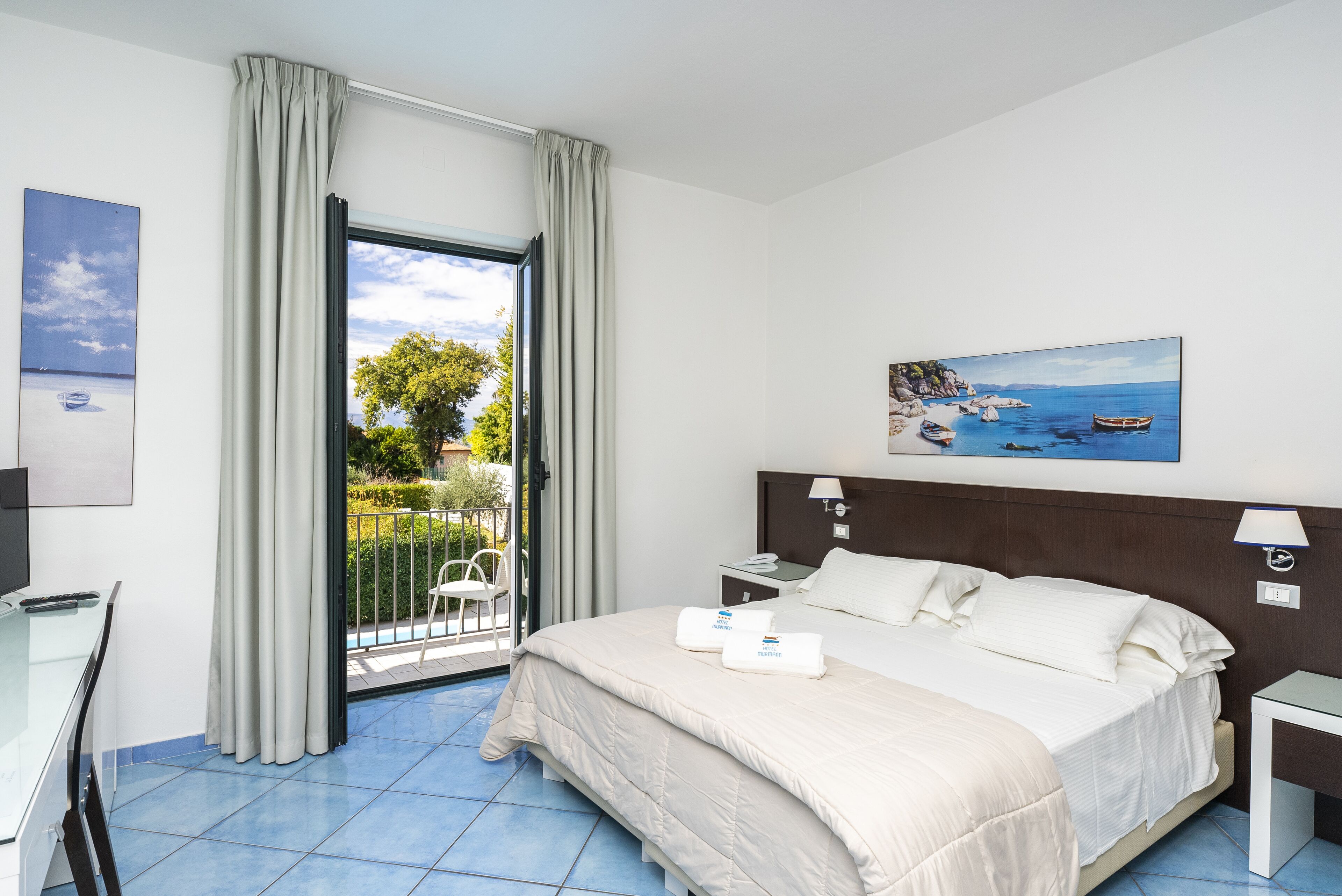 A hotel room with a bed, a desk, a chair, and a balcony with a view of the sea.