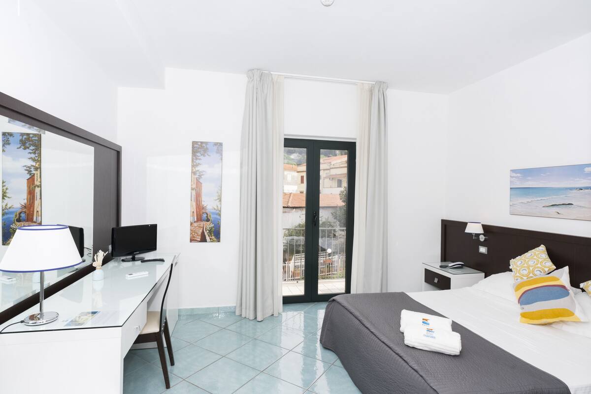 A hotel room with a desk, bed, and a balcony with a view of buildings.
