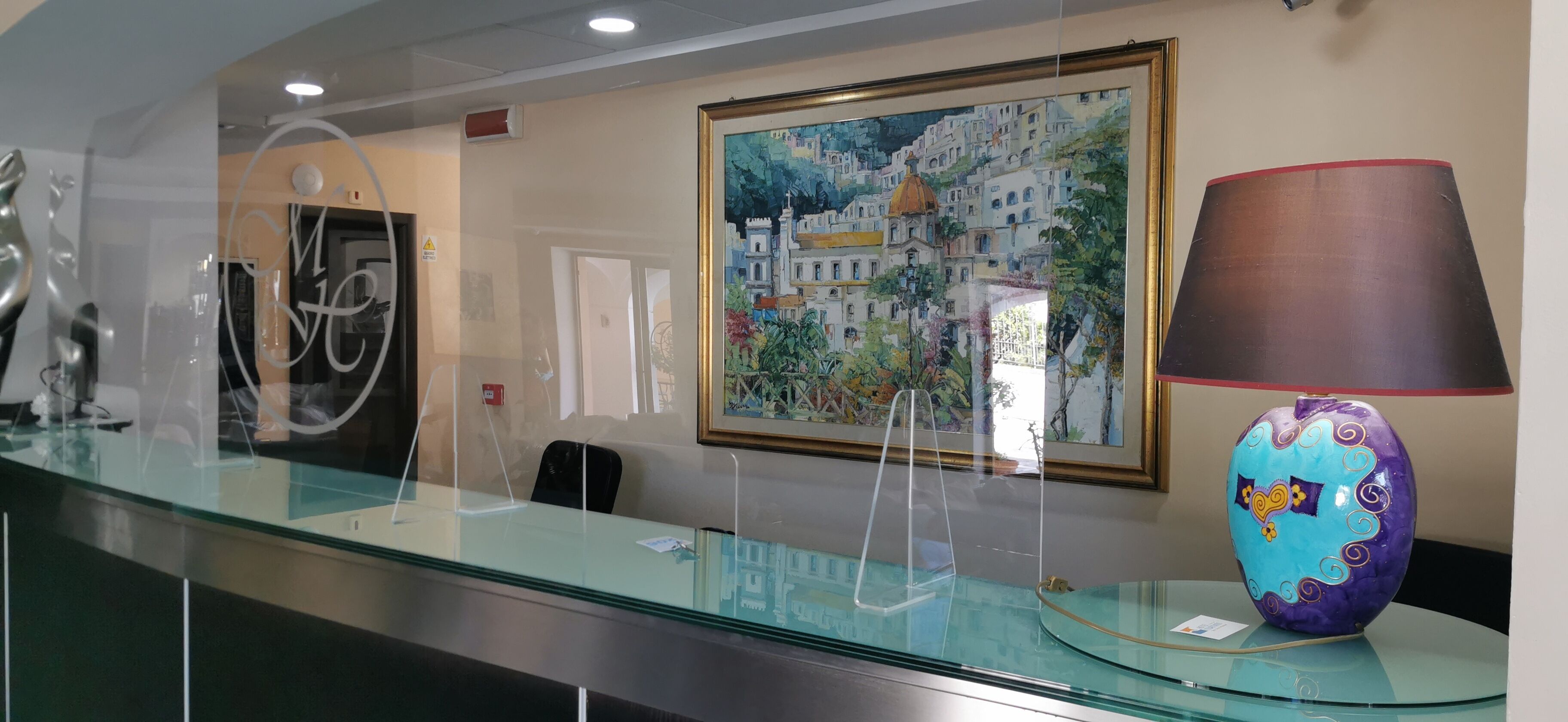 A reception desk with a glass top, a framed painting of a coastal town, a decorative lamp, and a glass vase.