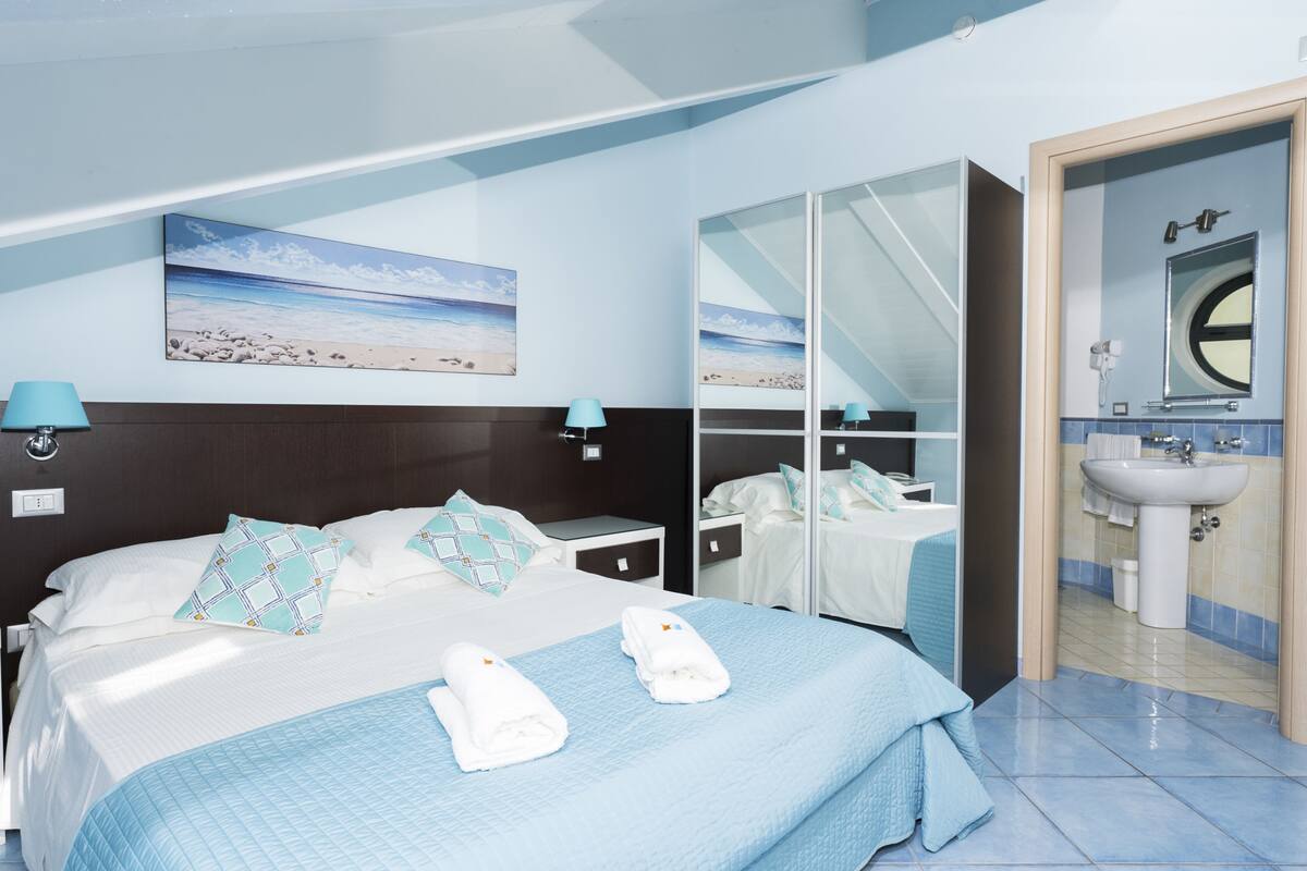 A hotel room with a bed, a large mirror, a bathroom with a sink, and a beach scene on the wall.