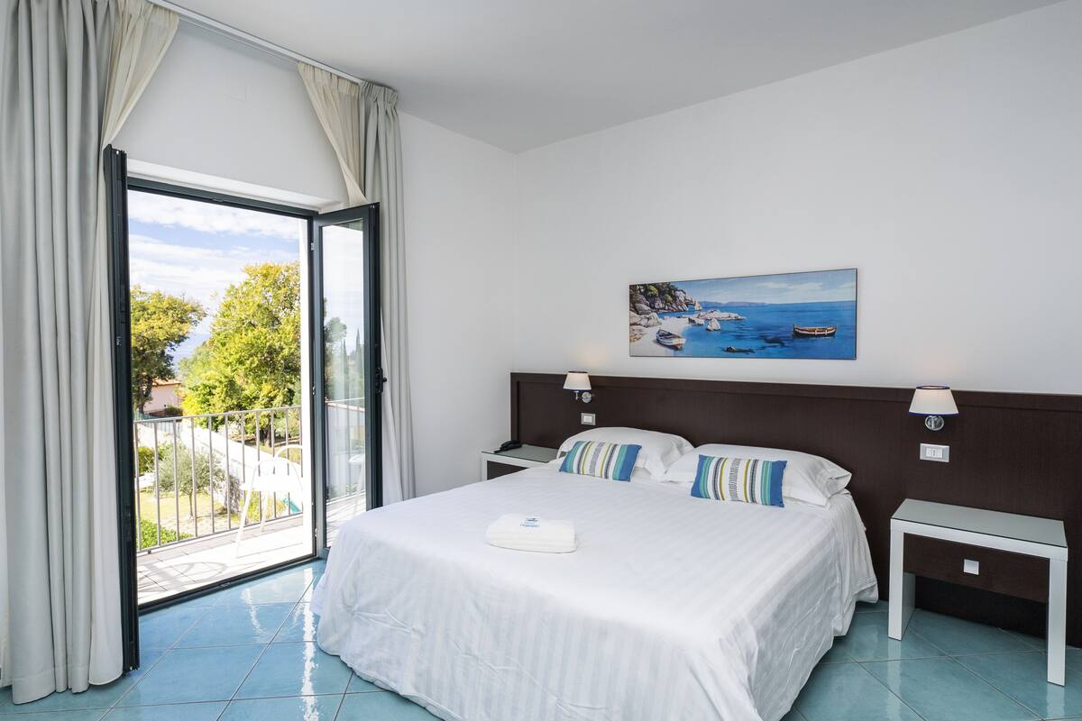 A hotel room with a large bed, a painting of a coastal scene, a balcony with curtains, and a view of the outdoors.