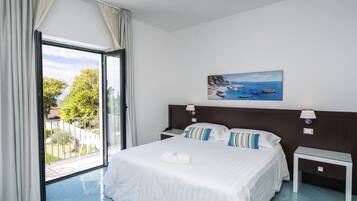 A hotel room with a large bed, a painting of a coastal scene, a balcony with curtains, and a view of the outdoors.