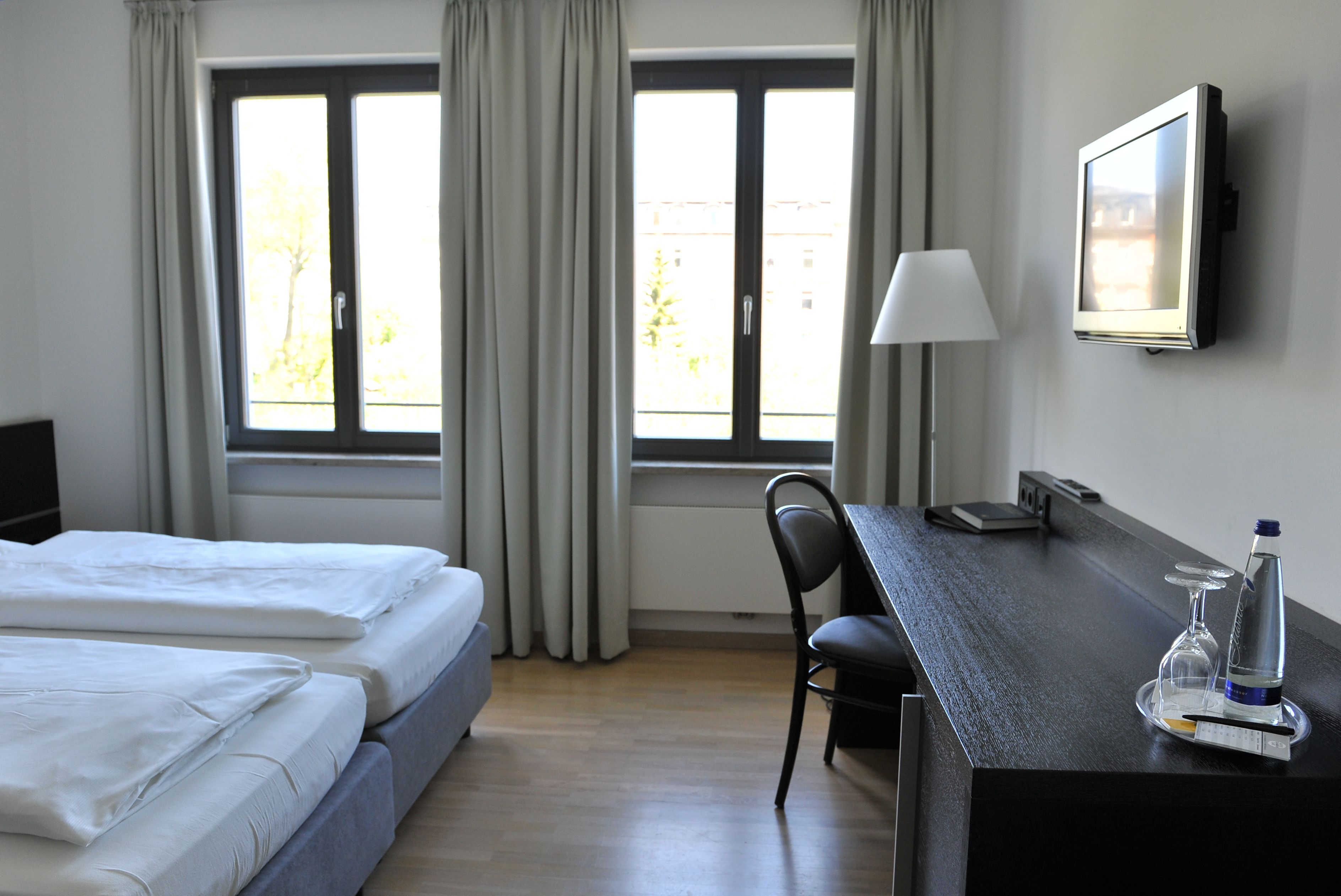 Habitación de hotel con cama, escritorio, silla, televisor y ventana con cortinas.