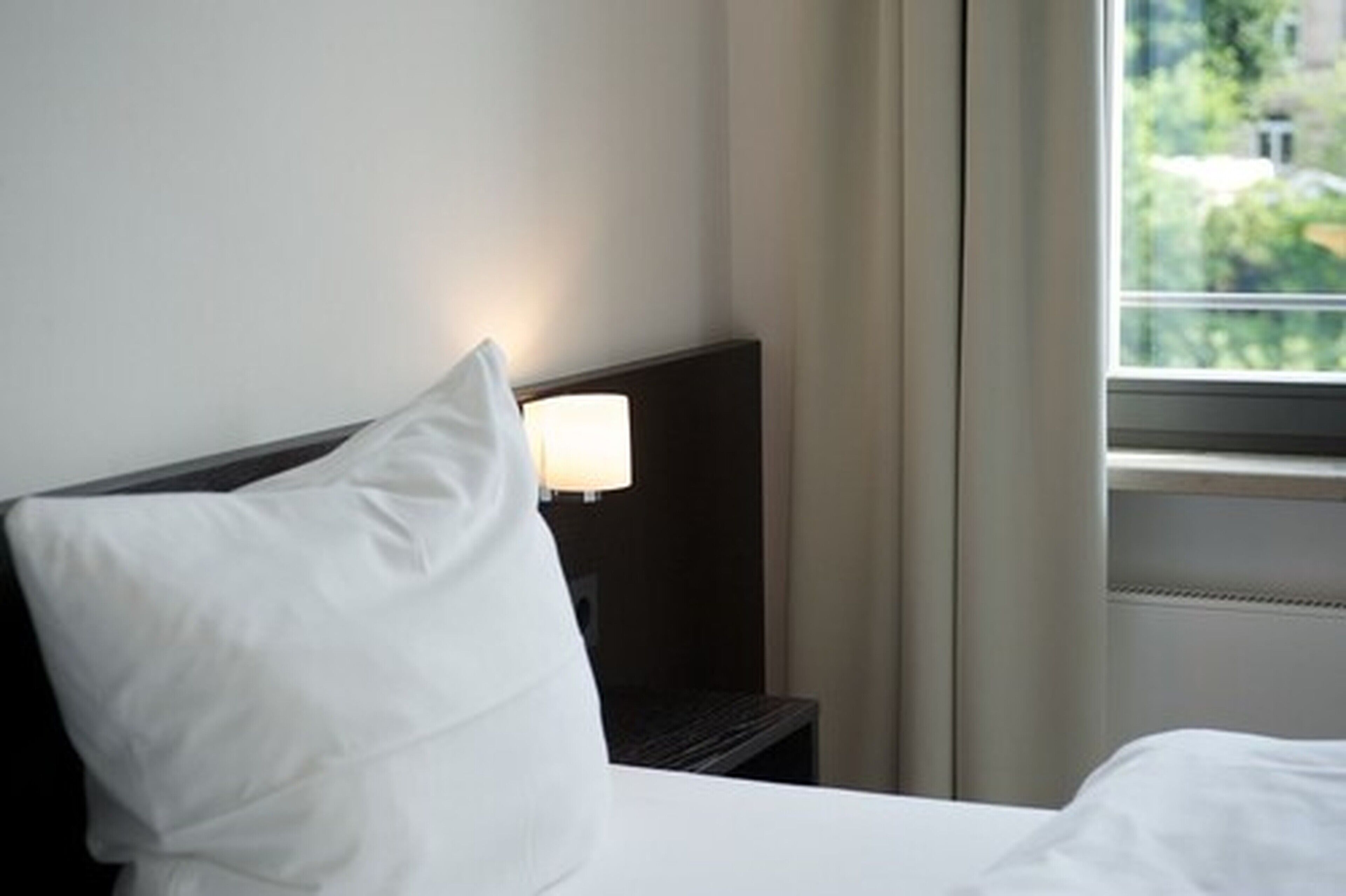 A hotel room with a bed, a nightstand with a lamp, and a window with a view of greenery.