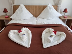 Ein ordentlich bezogenes Bett mit weißen Leinentüchern, zwei Kissen, einer braunen Decke und zwei mit roten Blumen verzierten Handtüchern, die auf dem Bett gefaltet liegen.