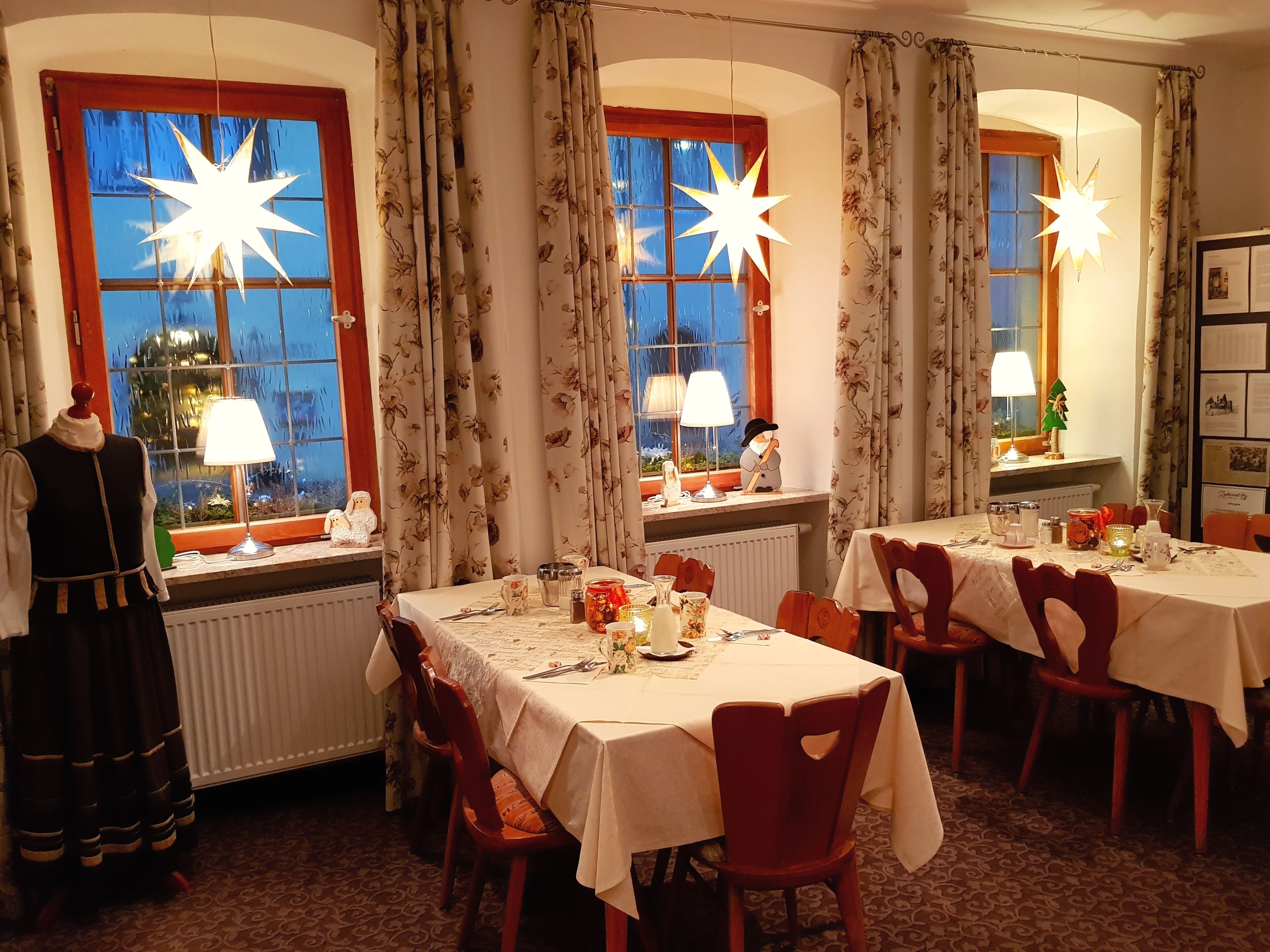 Ein Essbereich mit vier gedeckten Tischen, jeder Tisch ist mit einer weißen Tischdecke und floralen Mittelstücken dekoriert. Drei Fenster mit floralen Vorhängen und sternförmige Leuchten darüber. Eine Puppa in traditioneller Tracht steht links.