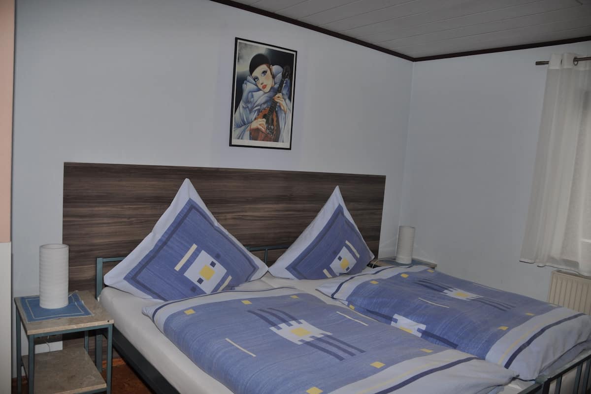 Ein Doppelbett mit Holz-Kopfteil, zwei Kissen, ein Nachttisch mit Lampe und ein gerahmtes Bild an der Wand.