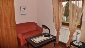 Ein Zimmer mit einer roten Ledersofa, einem kleinen Tisch mit Lampe, einem Heizkörper und durchsichtigen Vorhängen vor einem Fenster.