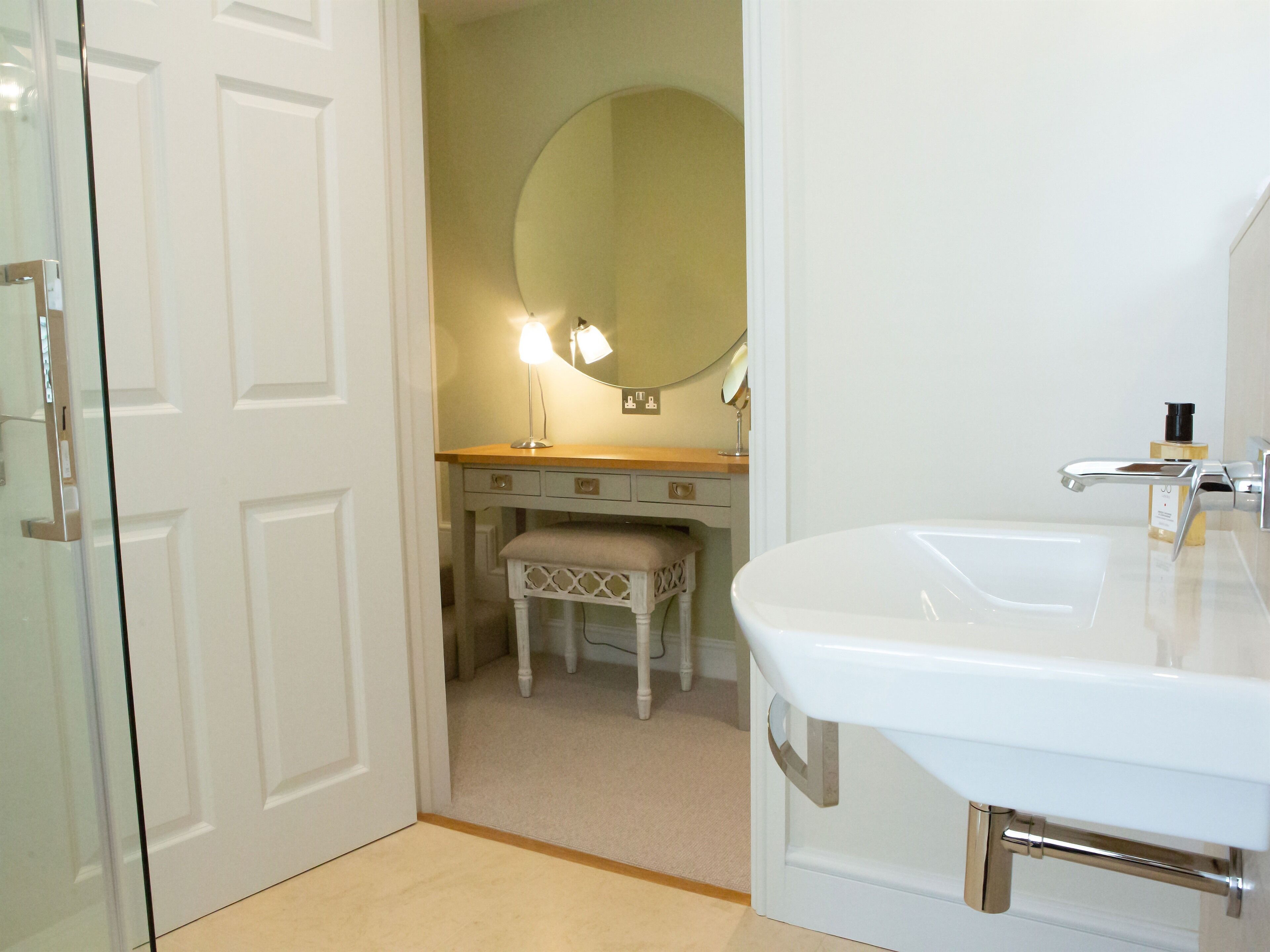 Un baño con un lavamanos blanco, un espejo, una tocador con una lámpara y una puerta que da a otra habitación.