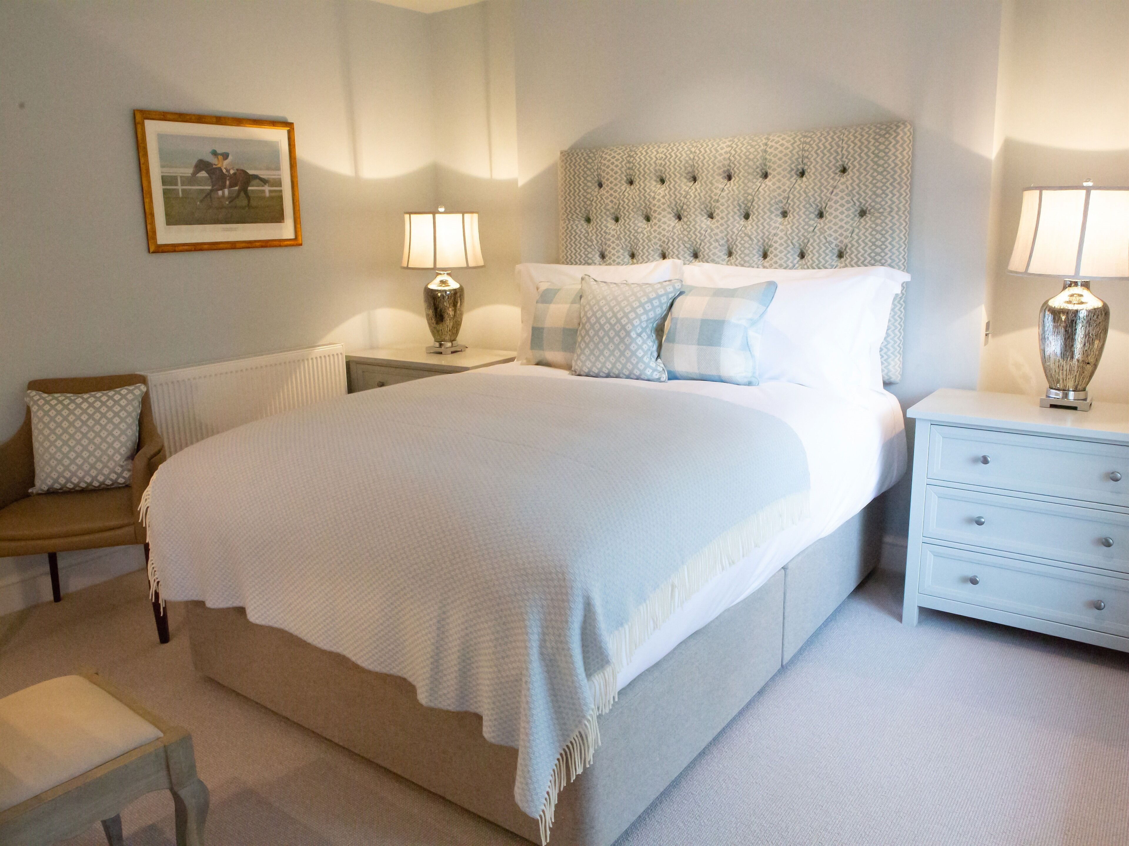 A neatly arranged bedroom with a large bed, a bedside table with lamps, a chair, and a framed picture on the wall.