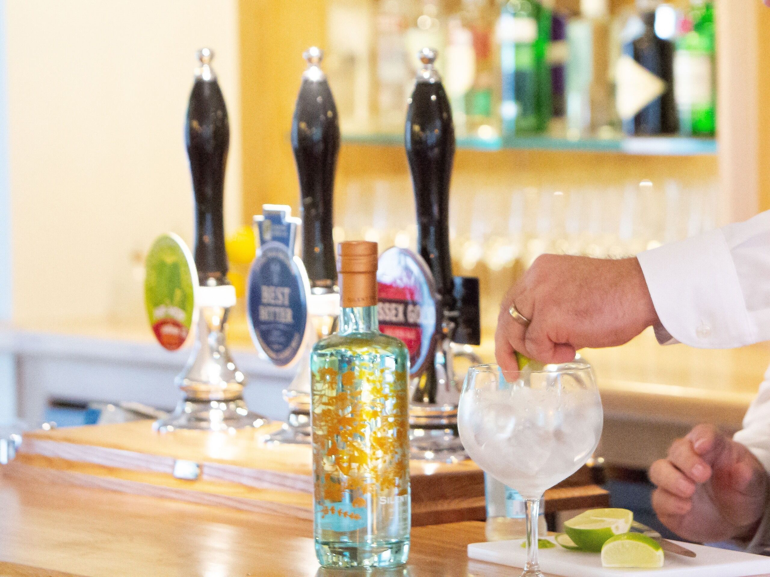 Een bartender giet een drankje in een glas achter een bar met verschillende biertaps en een fles drank.