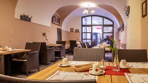 A cozy dining area with wicker furniture, tables set with white cups and a red placemat, and a large window offering a view of the street. - Pension U Lilie (Prague)