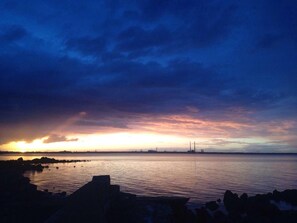 A sunset over a calm body of water with a rocky shoreline and distant industrial structures. - Acara House (Dublin)